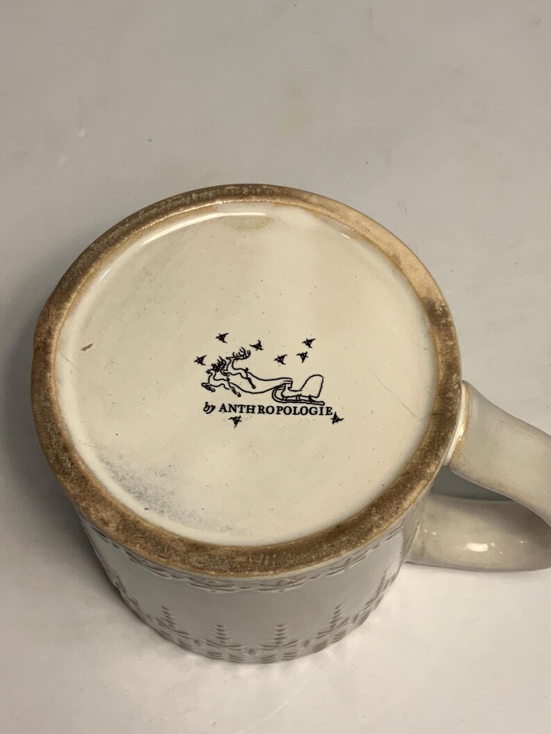 Peut inclure: Mug en c&eacute;ramique cr&egrave;me avec un motif textur&eacute; et une anse incurv&eacute;e. Le fond du mug pr&eacute;sente une illustration noire d'un renne tirant un tra&icirc;neau, avec le texte "by ANTHROPOLOGIE".