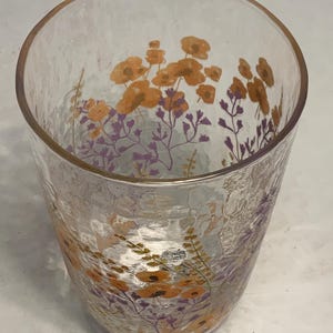 May include: A clear glass tumbler with a textured surface. The glass is decorated with orange and purple floral designs. The flowers are clustered near the top of the glass, creating a decorative pattern.