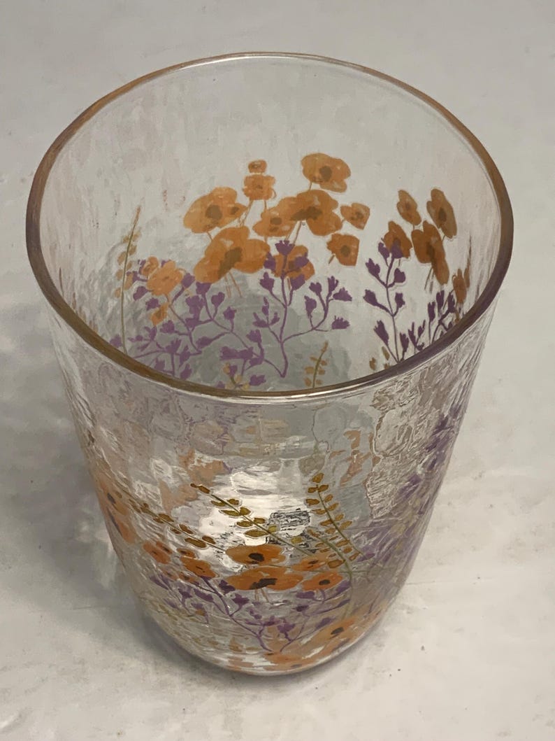 May include: A clear glass tumbler with a textured surface. The glass is decorated with orange and purple floral designs. The flowers are clustered near the top of the glass, creating a decorative pattern.