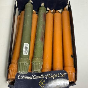 May include: A box of Colonial Candle of Cape Cod taper candles in shades of olive green and burnt orange. The candles feature a fluted base design and a white wick at the top. The dark blue packaging displays the brand name and a gold logo.