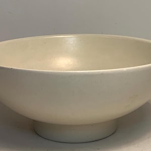 Könnte beinhalten: Eine cremefarbene Keramikschale mit runder Form und Sockel. Die Schale hat eine glatte, matte Oberfläche und eignet sich zum Servieren von Speisen oder als dekoratives Objekt. Der Durchmesser der Schale beträgt ca. 25 cm.