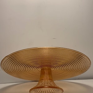 May include: A vintage amber-colored glass cake stand. The stand features a circular, ridged plate supported by a pedestal base. The plate has concentric ridges, and the base has vertical ridges. The cake stand is a decorative serving piece.