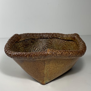 May include: A square, handmade ceramic bowl with a textured, brown exterior. The rim features a decorative rope-like design. The interior has a sunburst pattern. The bowl has a rustic, earthy aesthetic.