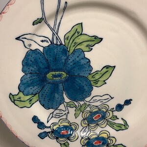 May include: White ceramic plate with a blue floral design. The plate features a large blue flower with green leaves and smaller blue and yellow flowers. The rim has a red and white scalloped border. Decorative tableware.