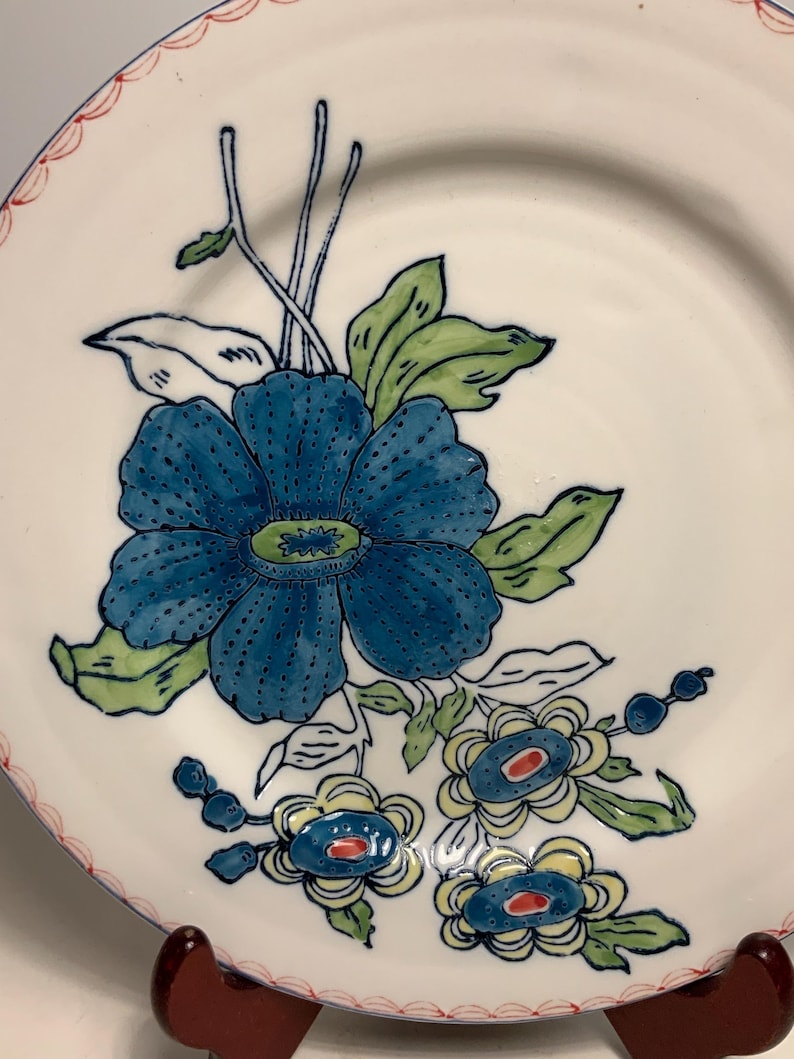 May include: White ceramic plate with a blue floral design. The plate features a large blue flower with green leaves and smaller blue and yellow flowers. The rim has a red and white scalloped border. Decorative tableware.