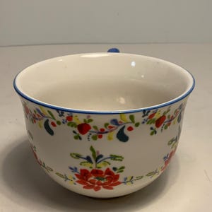 May include: White ceramic bowl with a blue rim and handle. The bowl is decorated with a floral pattern in red, yellow, and blue. The bowl is a round shape.