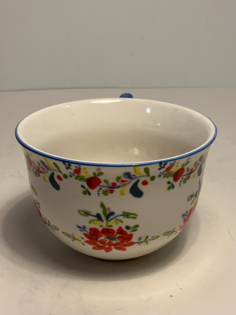 May include: White ceramic bowl with a blue rim and handle. The bowl is decorated with a floral pattern in red, yellow, and blue. The bowl is a round shape.
