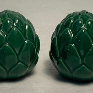 Op de afbeelding: Twee groene keramische zout- en peperstrooiers in de vorm van artisjokken. Elke strooier heeft een glanzende afwerking en kleine gaatjes aan de bovenkant om te doseren. De artisjokstrooiers zijn een keukenaccessoire.