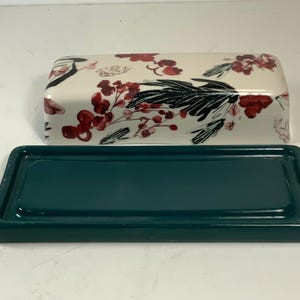 May include: A rectangular ceramic butter dish with a teal base and a lid. The lid is white with a floral pattern of red berries and black leaves. The dish is designed to hold butter.