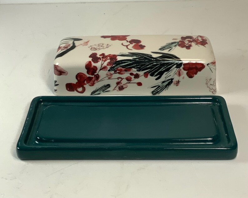 May include: A rectangular ceramic butter dish with a teal base and a lid. The lid is white with a floral pattern of red berries and black leaves. The dish is designed to hold butter.