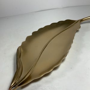 Puede incluir: Una bandeja de servir dorada en forma de hoja con un borde ondulado. La bandeja est&aacute; hecha de metal y tiene un acabado liso y brillante.