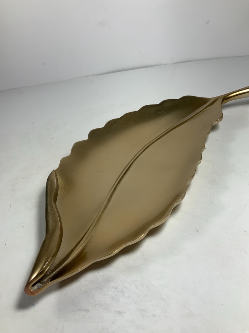 Puede incluir: Una bandeja de servir dorada en forma de hoja con un borde ondulado. La bandeja est&aacute; hecha de metal y tiene un acabado liso y brillante.