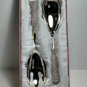 May include: A silver-toned salad serving set consisting of a fork and a spoon, presented in a pink box with white satin lining. Each utensil features a polished metallic finish and a handle encrusted with numerous small, clear, reflective stones.