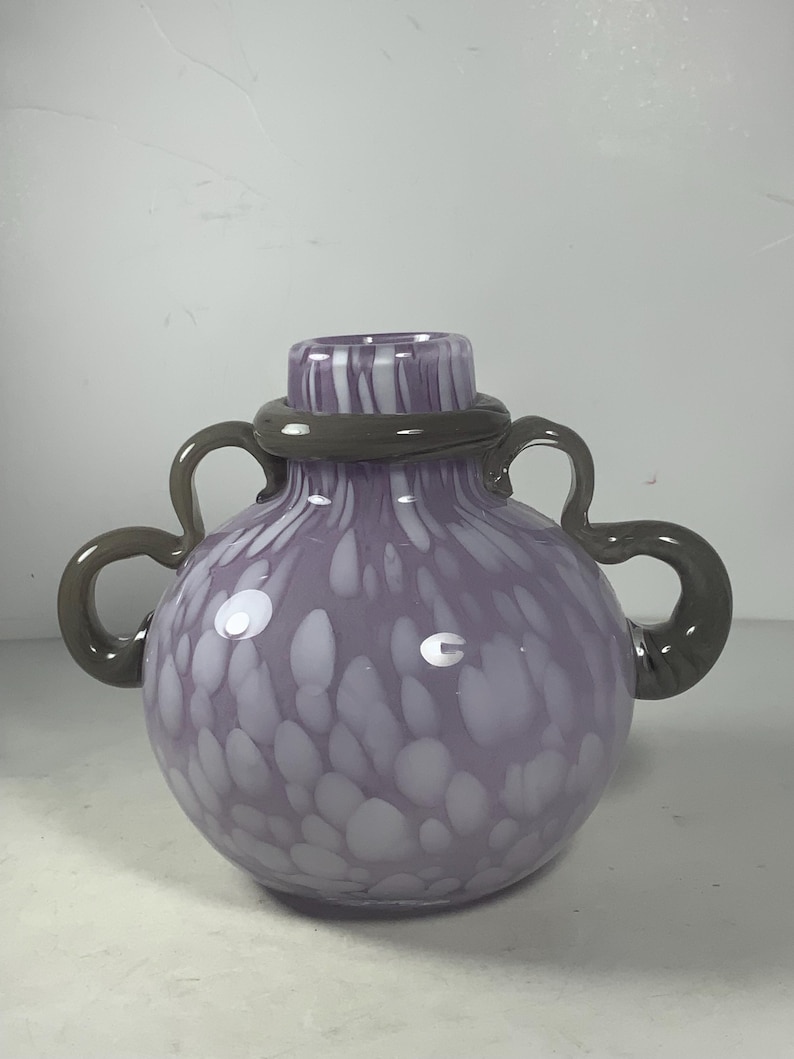 May include: A purple glass vase with white speckles and a grey swirl around the neck. The vase has two grey handles.