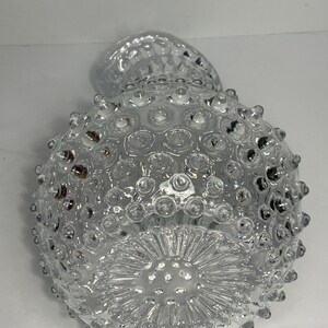 May include: Clear glass vase with a textured, hobnail design. The vase has a round body and a flared base with a starburst pattern. The top of the vase has a small, rounded opening.