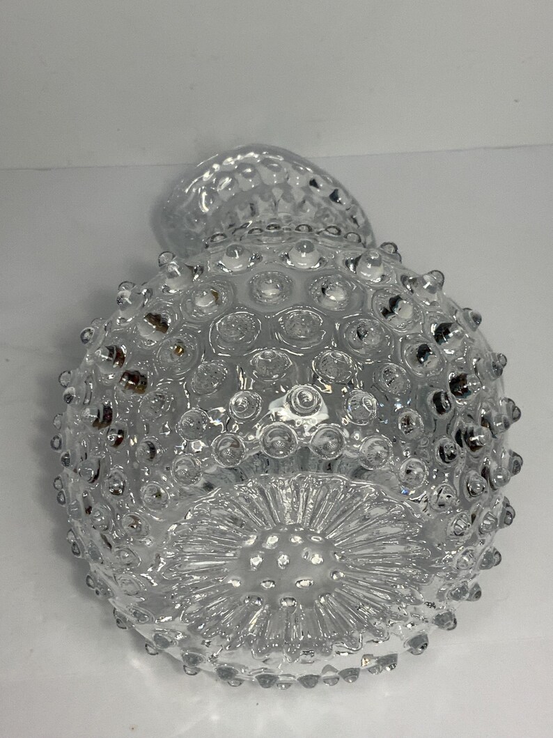 May include: Clear glass vase with a textured, hobnail design. The vase has a round body and a flared base with a starburst pattern. The top of the vase has a small, rounded opening.