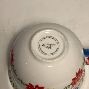 May include: White ceramic bowl with a floral pattern in red, yellow, and blue. The bowl has a blue handle and a circular stamp on the bottom that reads "Microwave & Dishwasher Safe" and "Made in China".