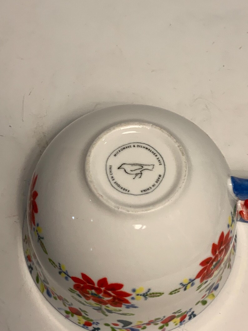 May include: White ceramic bowl with a floral pattern in red, yellow, and blue. The bowl has a blue handle and a circular stamp on the bottom that reads "Microwave & Dishwasher Safe" and "Made in China".