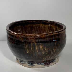 May include: A handcrafted ceramic bowl with a glossy, dark brown glaze. The bowl features a rounded shape with a slightly flared rim and a small, circular base. The glaze has a dripped effect, creating a unique texture.