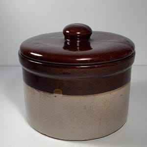 Peut inclure: Un pot en céramique bicolore avec un couvercle. La moitié supérieure du pot et le couvercle sont brun foncé, tandis que la moitié inférieure est beige clair. Le couvercle a un petit bouton arrondi.