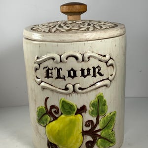May include: White ceramic flour canister with a brown wooden knob lid. The canister has a brown floral design on the lid and a brown pear with green leaves on the front. The word "Flour" is written in brown lettering on the front.