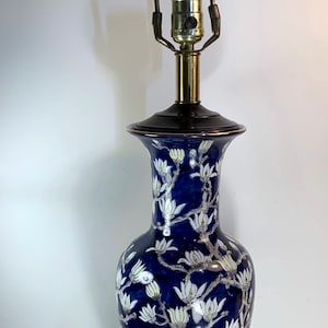 以下が含まれることがあります： 花柄の青いセラミックテーブルランプ。ランプは、白い花と緑の葉が付いた濃い青色のベースが特徴です。ランプは真鍮の首と装飾的なフィニアルが付いています。ランプの高さは約71cmです。