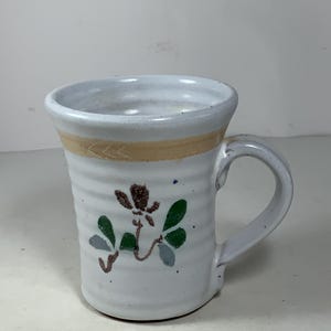 May include: White ceramic mug with a brown and green floral design. The mug has a light brown band around the top and a handle.