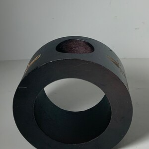 Puede incluir: Un anillo de madera negro con un peque&ntilde;o agujero en la parte superior. El anillo tiene un acabado liso y pulido y est&aacute; decorado con un dise&ntilde;o dorado.