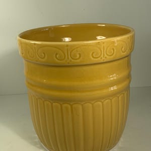 Puede incluir: Una maceta de cerámica amarilla con un borde decorativo y detalles acanalados verticales. La maceta tiene una parte superior ligeramente ensanchada y un acabado liso y brillante. El diseño incluye una banda de volutas cerca de la parte superior.