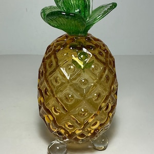 Puede incluir: Una figura decorativa de piña de cristal con un cuerpo texturizado de color ámbar y una superficie con patrón de diamantes. La parte superior consta de hojas de cristal verde translúcido y la base se apoya en tres pequeños pies de cristal transparente.