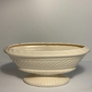 May include: Cream-colored ceramic planter with a pedestal base. The planter has a wide, shallow bowl shape with a textured, ridged design around the body. The base is a circular shape with a matching ridged pattern. A decorative piece for plants.