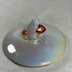 Może przedstawiać: Biała ceramiczna pokrywka z iryzującym wykończeniem. Mała ceramiczna figurka ptaka siedzącego na grzybie jest przymocowana do pokrywki.
