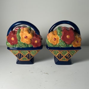 May include: A pair of ceramic salt and pepper shakers shaped like baskets overflowing with colourful flowers. The baskets are blue with gold accents and the flowers are red, yellow, and green.