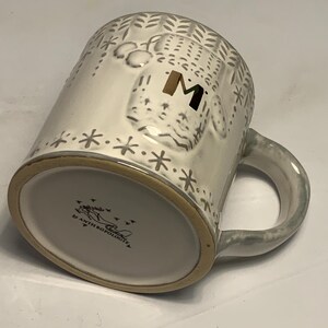 May include: A white ceramic mug with a gray handle and rim. The mug features a raised design of a bear with the letter "M" in gold. The mug is decorated with snowflakes and leaf patterns. The bottom of the mug has the text "ANTHROPOLOGIE".