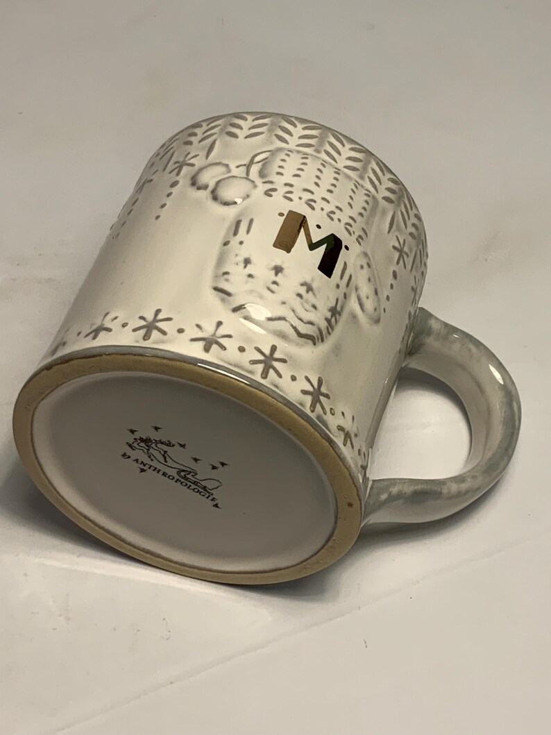 May include: A white ceramic mug with a gray handle and rim. The mug features a raised design of a bear with the letter "M" in gold. The mug is decorated with snowflakes and leaf patterns. The bottom of the mug has the text "ANTHROPOLOGIE".