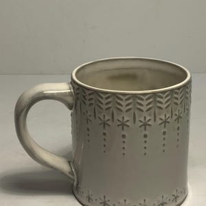 May include: A ceramic mug with a light gray glaze and a curved handle. The mug is decorated with a repeating pattern of stylized leaves and star-like shapes. The interior of the mug is a lighter shade of gray.
