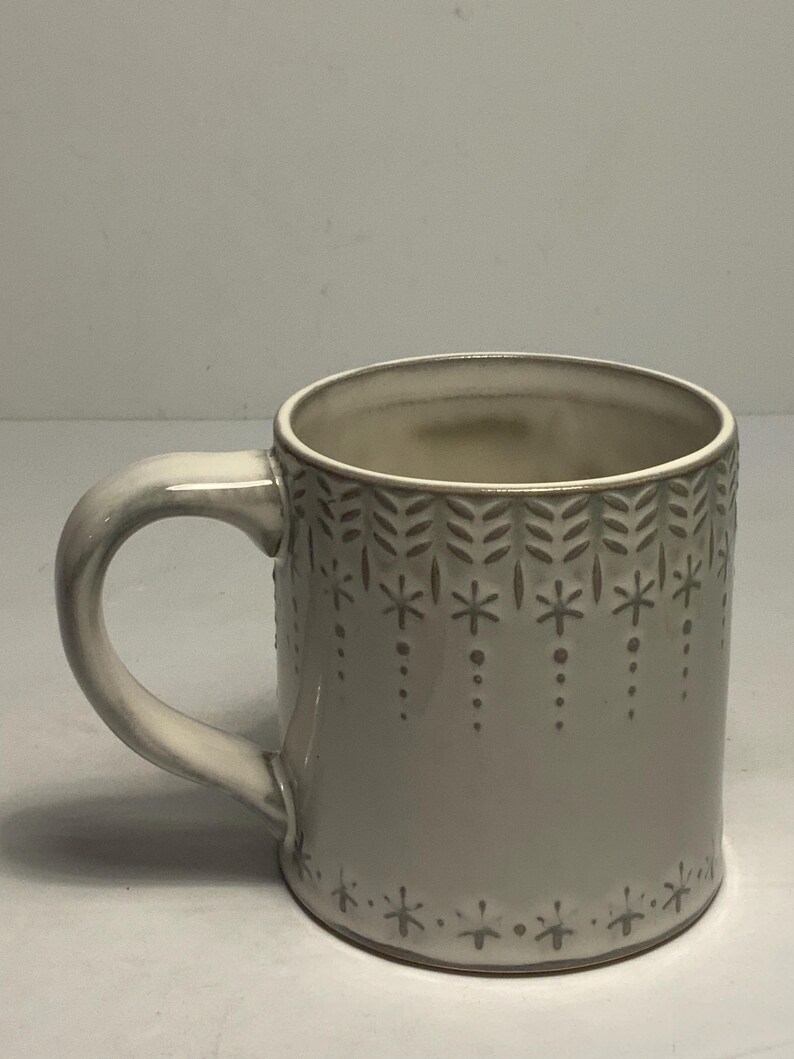 May include: A ceramic mug with a light gray glaze and a curved handle. The mug is decorated with a repeating pattern of stylized leaves and star-like shapes. The interior of the mug is a lighter shade of gray.