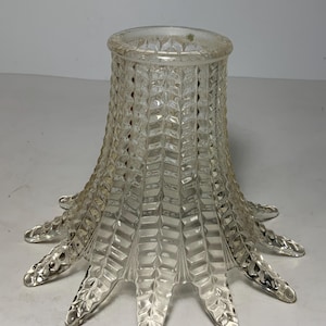 Peut inclure: Un abat-jour en verre transparent présentant un motif géométrique texturé et répétitif sur toute sa surface. L'abat-jour a une forme évasée en cloche avec un bord inférieur festonné en forme de pétale et une ouverture circulaire lisse.