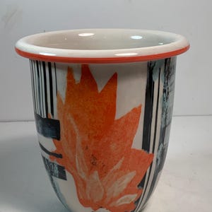 May include: A ceramic vase with a white base, featuring an orange floral design. The vase has a white rim with an orange border and black and blue vertical stripes. The vase is a decorative item.