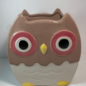 May include: A ceramic owl-shaped planter with a brown body, pink and white eyes, and a yellow beak. The bottom portion is white with a wavy design, and yellow accents are at the base. The planter has a wide opening at the top.