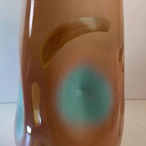May include: A tall, abstract art glass vase in shades of peach, brown, and teal. The vase has an organic shape with a wide base and a slightly narrower top. The colors blend and swirl, creating a unique decorative piece.