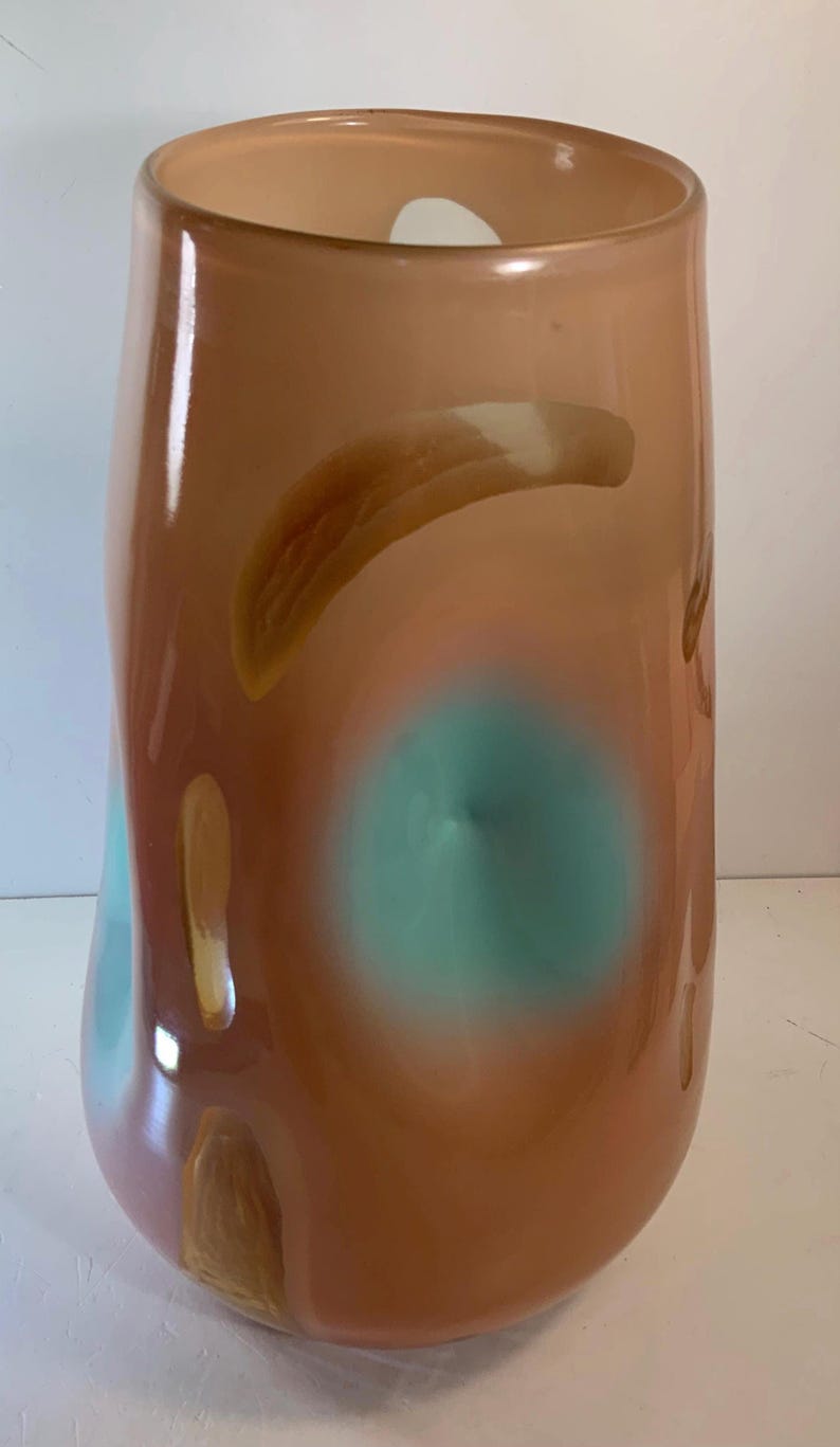 May include: A tall, abstract art glass vase in shades of peach, brown, and teal. The vase has an organic shape with a wide base and a slightly narrower top. The colors blend and swirl, creating a unique decorative piece.