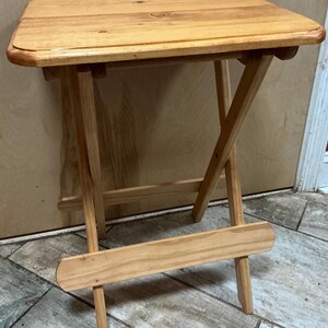 May include: A light brown wooden folding table with a rectangular top and rounded edges. The table features a cross-leg design with a horizontal support bar near the bottom, showcasing a natural wood grain finish throughout the entire structure.
