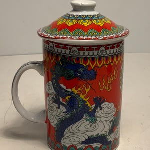 Può includere: Una tazza da tè in ceramica rossa con coperchio, con un disegno di drago blu. La tazza ha un manico bianco ed è decorata con motivi asiatici tradizionali in giallo, verde e blu. Il coperchio ha un pomello bianco.