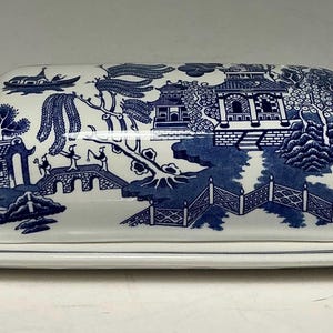 Op de afbeelding: Een witte keramische botervloot met een blauw wilgenpatroon met een landschap met een brug, pagode en bomen. Het rechthoekige deksel rust op een bijpassende onderkant met blauw gedessineerde handvatten aan beide zijden.