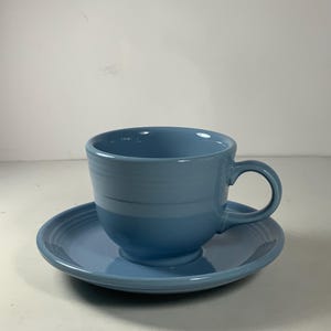 May include: A light blue ceramic teacup and saucer set. The cup has a rounded shape and a handle. The saucer has a slightly raised edge.