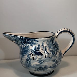May include: A ceramic pitcher with a blue and white landscape design. The pitcher features a spout, a handle, and a black base. The landscape depicts trees and buildings. The handle has a blue and white pattern.