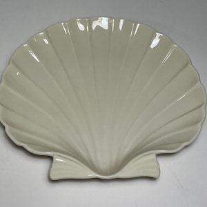 May include: A cream-colored ceramic dish shaped like a large scallop shell. The dish features a glossy finish with deep, radiating ridges that extend from the base to the scalloped outer edge, creating a textured, fan-like appearance.