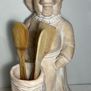 May include: A light brown ceramic figurine of a pig dressed as a chef, wearing a tall hat and apron. The pig stands behind a cylindrical container that holds two wooden spoons and a wooden spatula, all with visible natural wood grain textures.