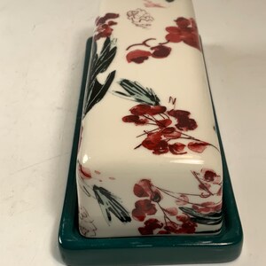 May include: A rectangular butter dish with a white lid featuring a floral design in red and black. The dish sits on a dark teal base. The floral pattern includes red flowers and black leaves.